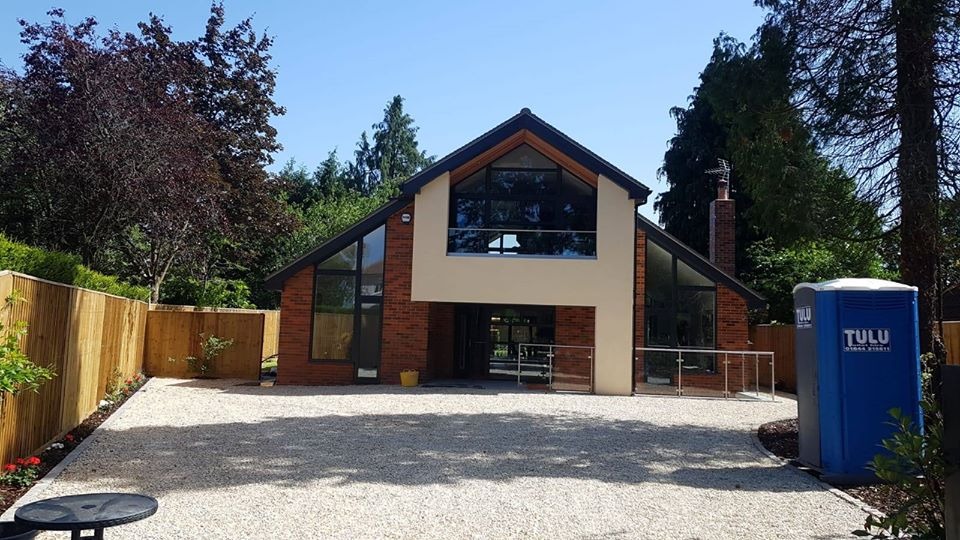 Nowoczesny dom z czerwonej cegły z dużymi oknami i szklanymi balustradami, jasny żwirowy podjazd, przenośna toaleta TULU z boku.