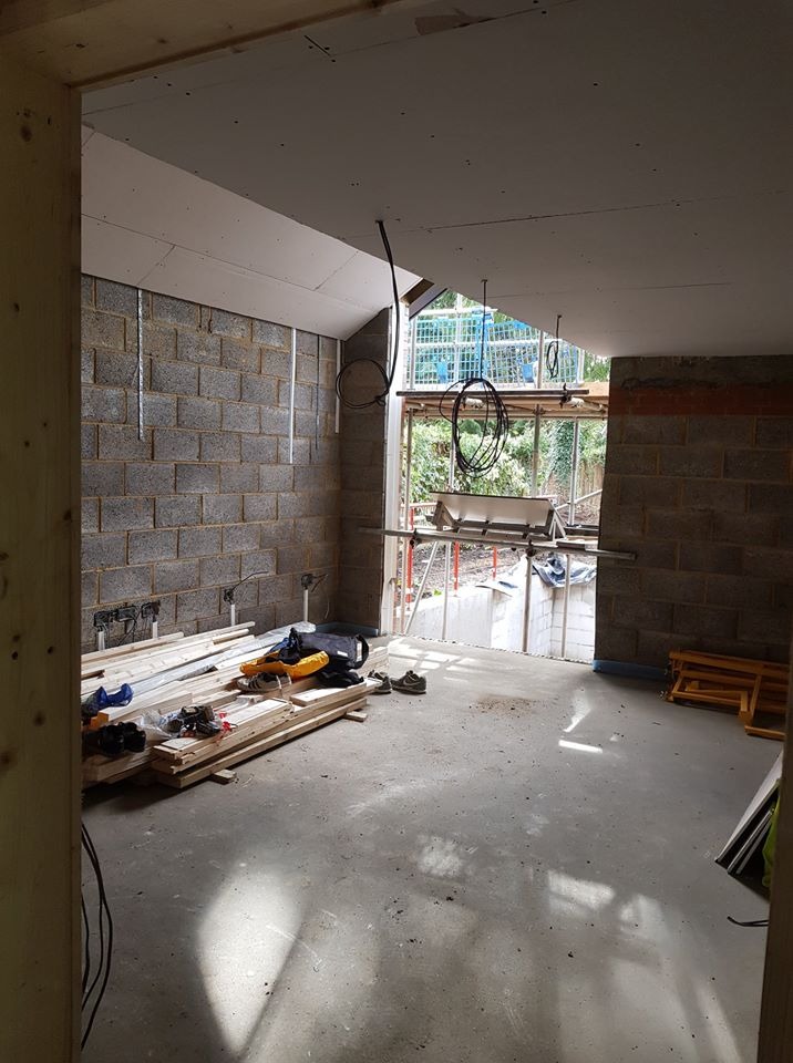 Wnętrze w trakcie remontu: surowe ściany z bloczków, częściowo otynkowany sufit, kable zwisające z sufitu, deski i narzędzia na podłodze, widok na zewnątrz przez otwór okienny z rusztowaniami.