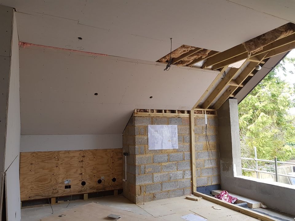 Wnętrze w trakcie remontu: surowy strop z widoczną izolacją, ściana z płyt OSB z puszkami elektrycznymi, fragment murowanej ściany z projektem, okno z widokiem na zieleń.