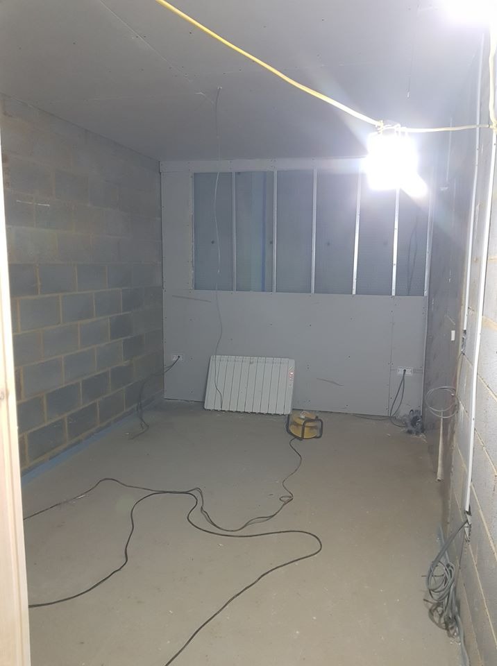 Wnętrze w trakcie remontu: surowe ściany z bloczków, częściowo zabudowana ściana działowa z metalowym stelażem i płytami gipsowymi, widoczne przewody elektryczne, grzejnik i lampa budowlana.