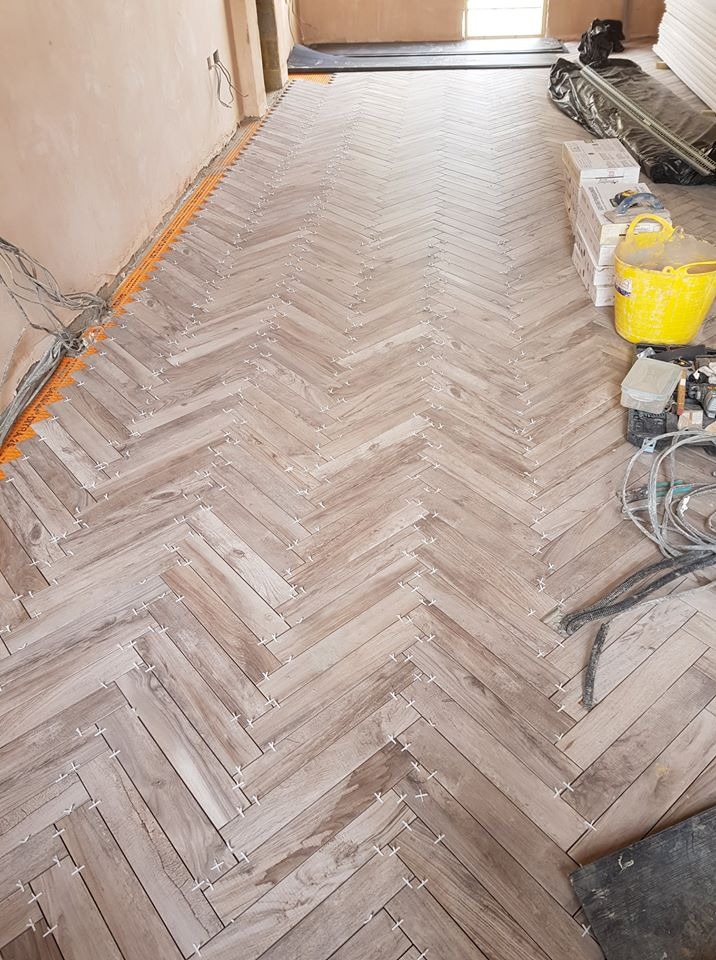 Układanie parkietu w jodełkę z użyciem białych krzyżyków dystansowych, widoczne narzędzia i materiały budowlane w tle, częściowo wykończona podłoga.