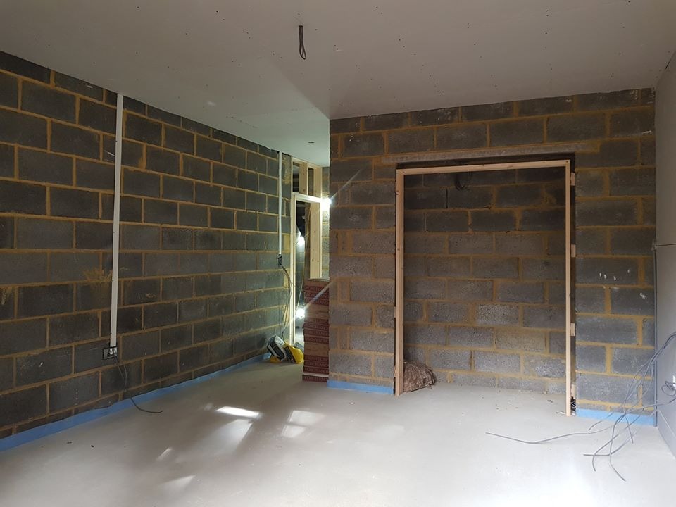 Surowe wnętrze w trakcie remontu: ściany z szarych bloczków, przygotowany otwór drzwiowy z drewnianą ramą, zabezpieczona podłoga, widoczne przewody elektryczne i narzędzia w tle.