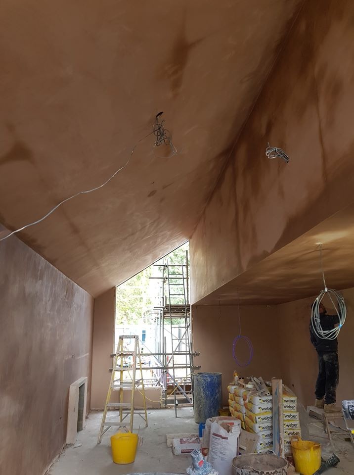 Wnętrze w trakcie tynkowania ścian i sufitu, widoczne rusztowania, worki z zaprawą Weber, wystające kable elektryczne i otwór okienny z widokiem na zieleń.