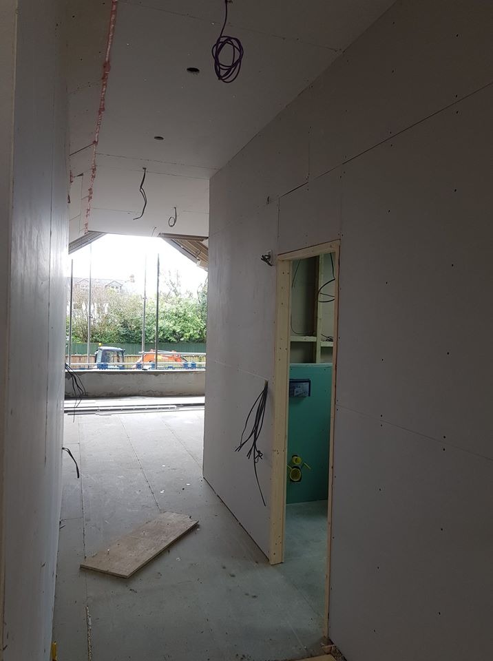 Wnętrze w trakcie remontu: surowe ściany z płyt gipsowo-kartonowych, wystające przewody elektryczne, widok na zewnątrz przez niezabudowane okno z rusztowaniem i sprzętem budowlanym, fragment...