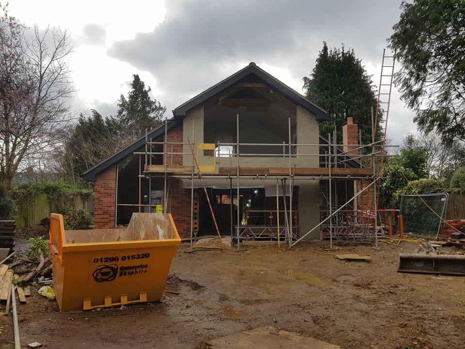 Dom w trakcie rozbudowy z widocznym rusztowaniem, kontenerem na gruz i elementami murarskimi, pochmurne niebo w tle.