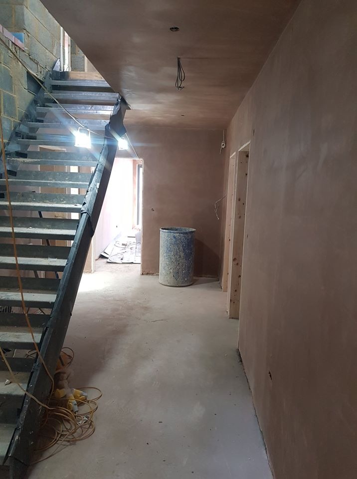 Wnętrze w trakcie remontu: ściany pokryte świeżym tynkiem, metalowe schody bez balustrady, niebieski plastikowy pojemnik, przewody elektryczne zwisające z sufitu, surowe ościeżnice drzwiowe.