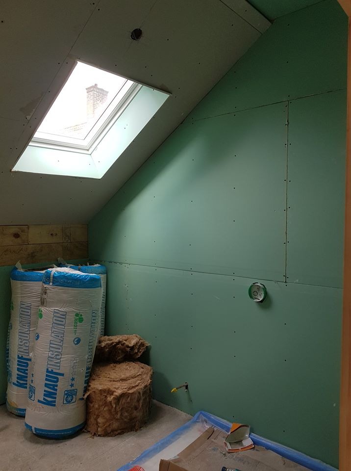 Poddasze w trakcie wykańczania: widoczne płyty gipsowo-kartonowe na ścianach, okno dachowe z widokiem na komin, rolki wełny mineralnej Knauf Insulation i fragment instalacji elektrycznej.