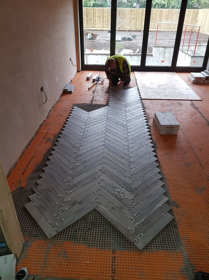 Układanie szarej podłogi z płytek w jodełkę na pomarańczowej macie izolacyjnej, widoczny pracownik w odblaskowej kamizelce, okno z widokiem na ogród.