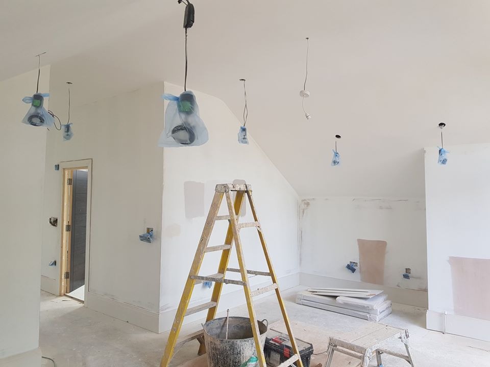 Pokój w trakcie malowania: widoczne zabezpieczone folią lampy wiszące, drabina, wiadro z farbą, szpachlowane ściany i podłoga z widocznymi śladami farby.