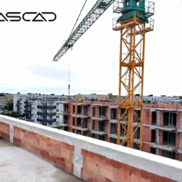 Budowa wielopiętrowego budynku z czerwonej cegły ceramicznej, widok z góry na konstrukcję w stanie surowym otwartym, z żurawiem budowlanym i logo firmy KASCAD w lewym górnym rogu.