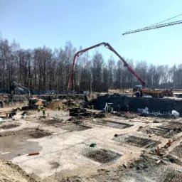 Budowa fundamentów pod budynek wielorodzinny: widok na plac budowy z szalunkami, zbrojeniami, pompą do betonu i pracującymi robotnikami w kamizelkach odblaskowych, na tle lasu i błękitnego nieba.