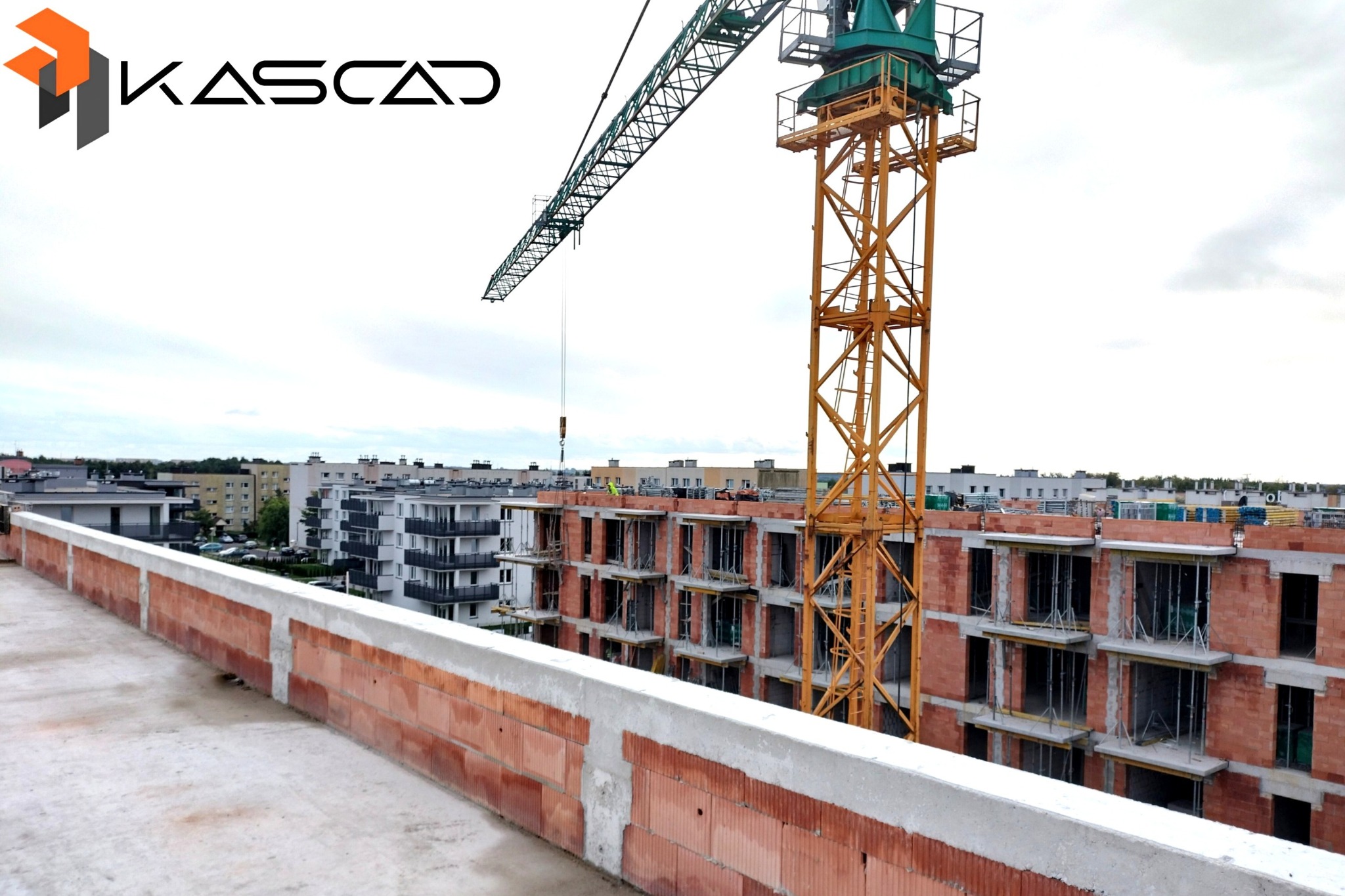 Budowa wielopiętrowego budynku z czerwonej cegły ceramicznej, widok z góry na konstrukcję w stanie surowym otwartym, z żurawiem budowlanym i logo firmy KASCAD w lewym górnym rogu.
