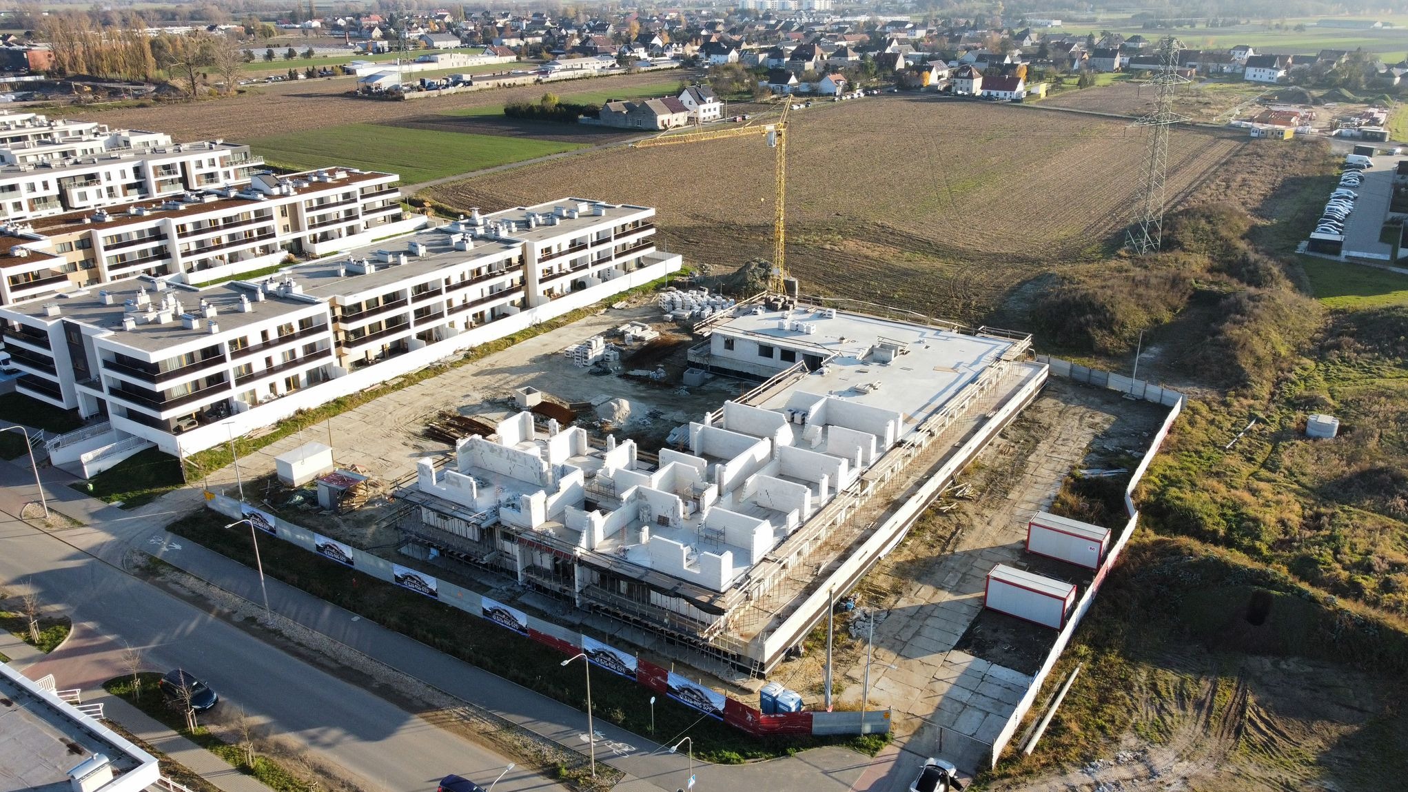 Widok z lotu ptaka na plac budowy wielorodzinnego budynku mieszkalnego w stanie surowym, z widocznymi ścianami działowymi i stropami, otoczonego ogrodzeniem i z żółtym dźwigiem w tle oraz nowym...