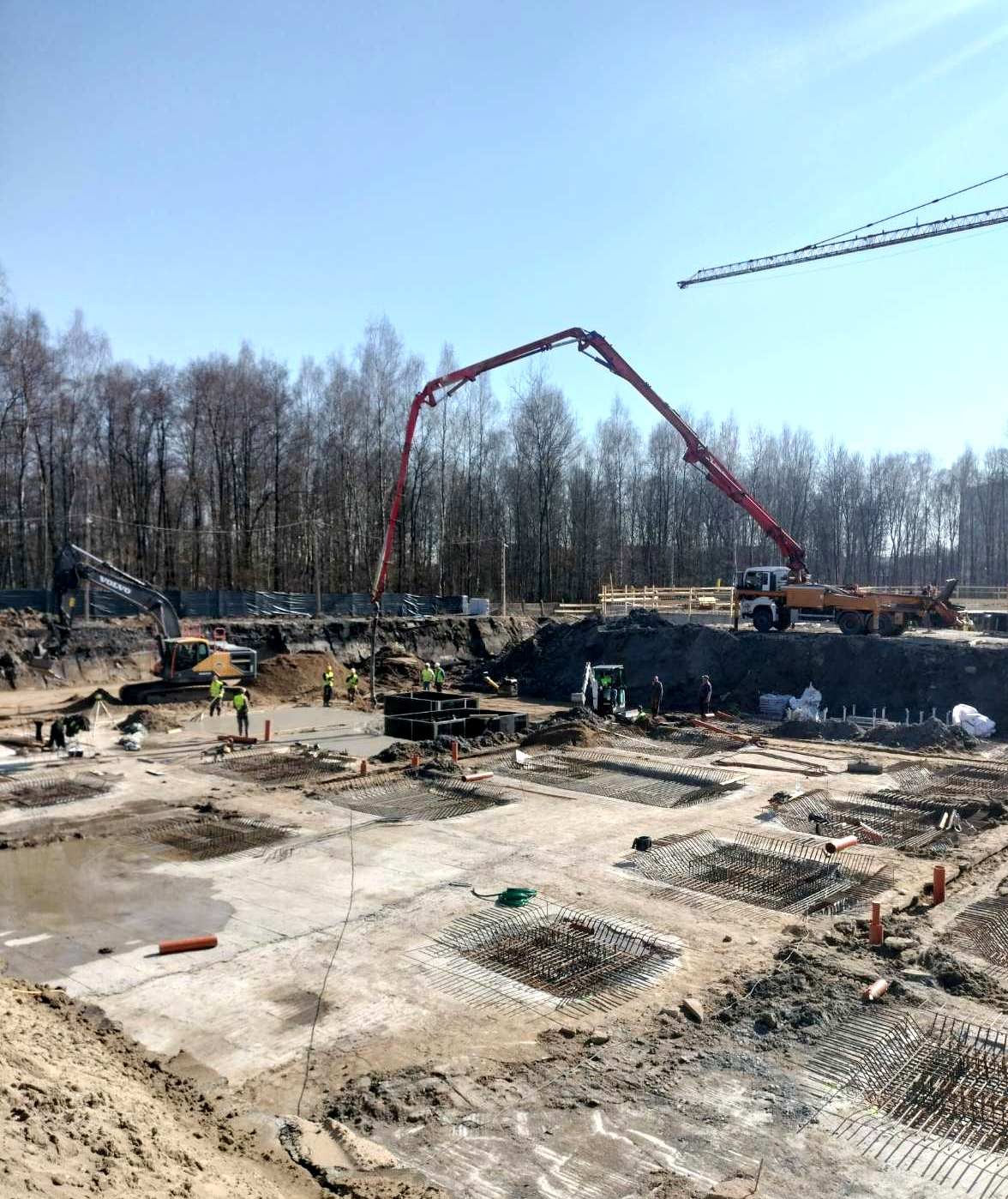 Budowa fundamentów pod budynek wielorodzinny: widok na plac budowy z szalunkami, zbrojeniami, pompą do betonu i pracującymi robotnikami w kamizelkach odblaskowych, na tle lasu i błękitnego nieba.