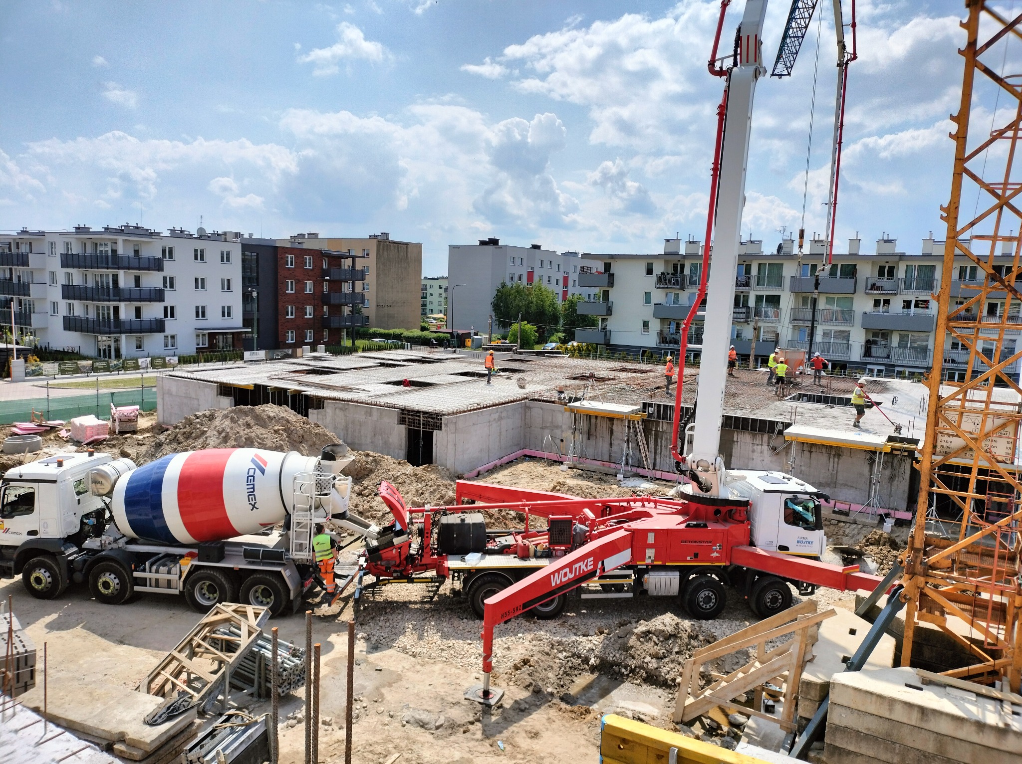 Budowa budynku wielorodzinnego w trakcie wylewania fundamentów i stropu, widoczny mikser do betonu Cemex, pompa do betonu Wojtke oraz żuraw wieżowy, w tle stoją gotowe bloki mieszkalne.