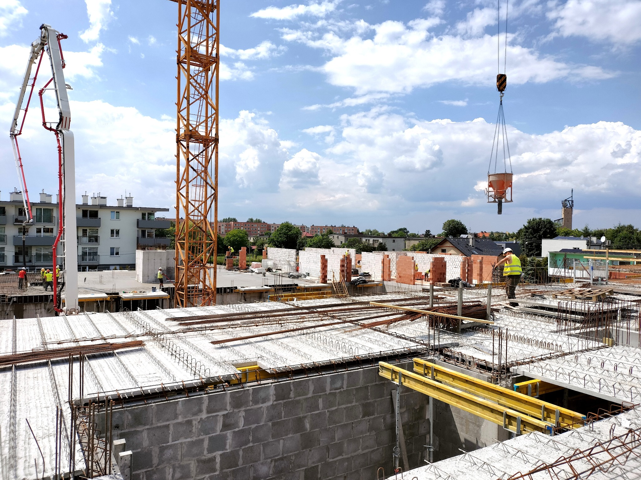 Budowa budynku wielorodzinnego w Opolu: widok z góry na zbrojenie stropu, częściowo wymurowane ściany z betonu komórkowego, dźwig podający beton i pracujących robotników w kamizelkach odblaskowych.