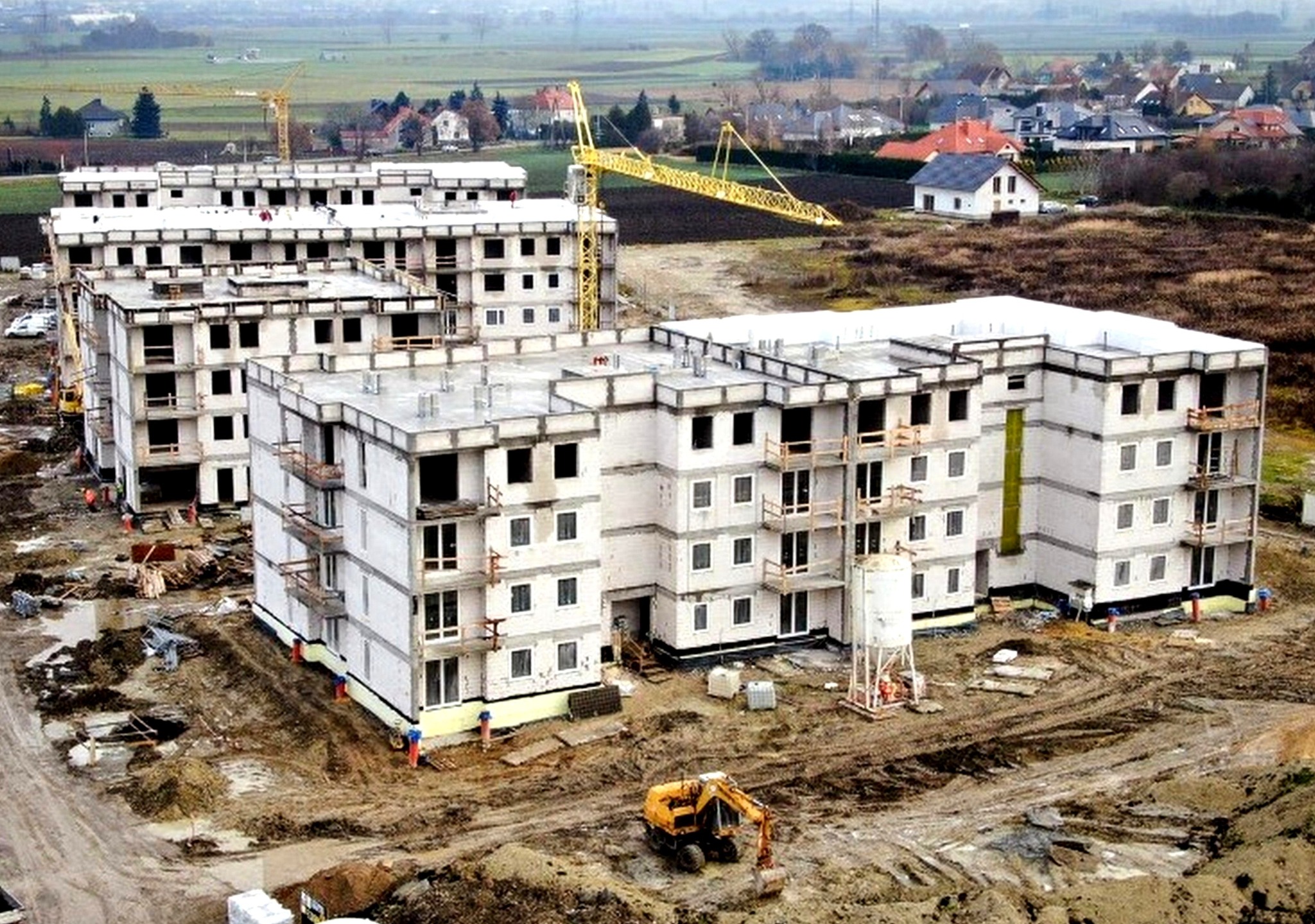 Wielorodzinny budynek mieszkalny w stanie surowym, z widocznym żelbetowym szkieletem i ścianami z betonu komórkowego, otoczony placem budowy z koparką i żurawiem wieżowym w tle.