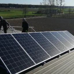 Montaż paneli słonecznych na metalowym dachu z widokiem na pola uprawne w tle, dwaj instalatorzy w roboczych ubraniach na dachu.