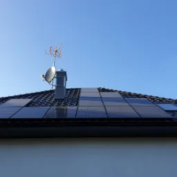 Panele słoneczne na ciemnym dachu z dachówkami, widoczne okna dachowe, antena satelitarna i komin na tle błękitnego nieba.