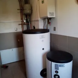 Biały, cylindryczny zasobnik ciepłej wody użytkowej z czarną pokrywą, obok mniejszy zasobnik, na ścianie wiszący piec gazowy kondensacyjny i instalacja rurowa