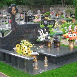 Granitowy pomnik nagrobny z płaskorzeźbą postaci, otoczony kwiatami i lampionami na cmentarzu, z widocznymi innymi nagrobkami w tle.