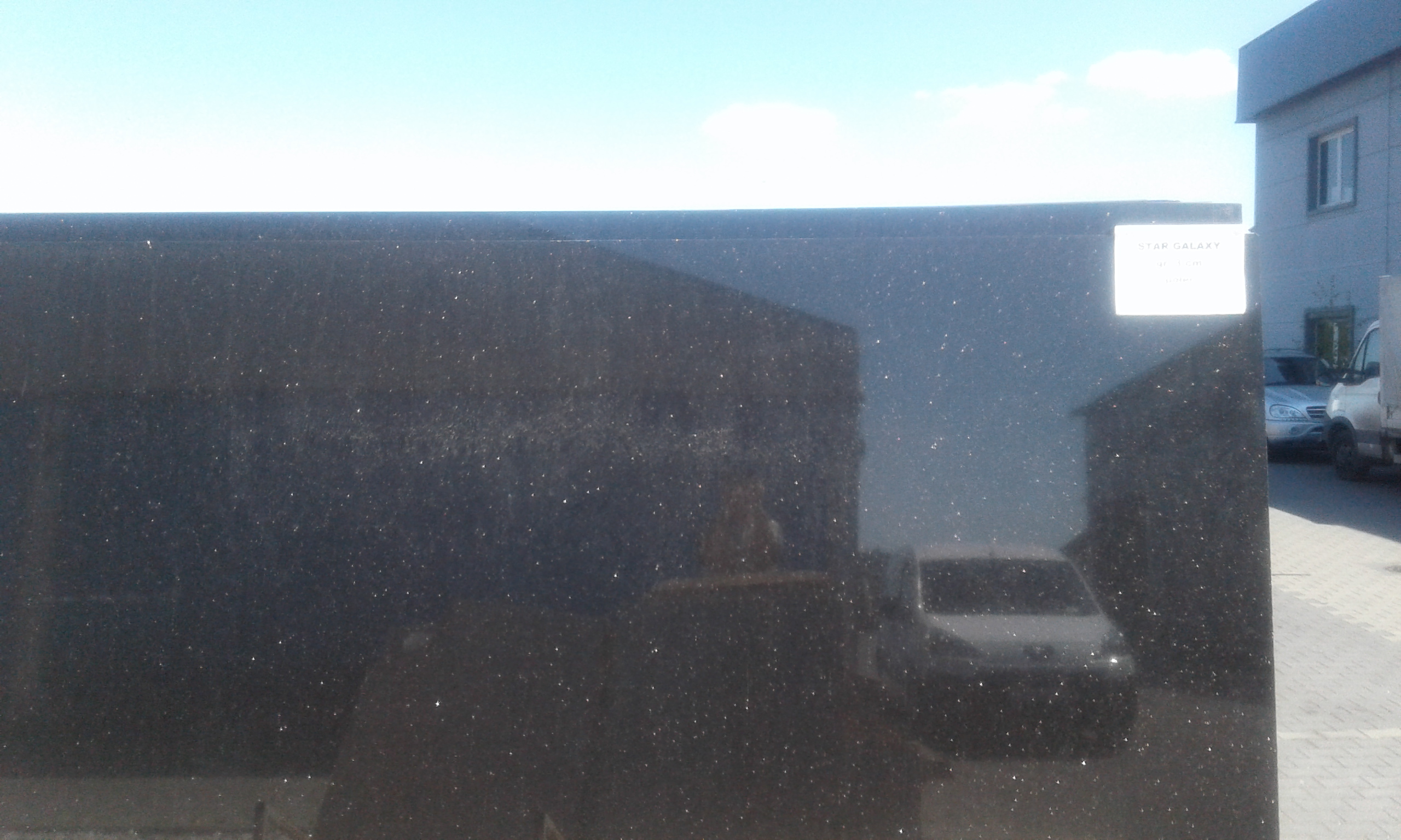 Ciemny, polerowany slab granitu Star Galaxy z naklejoną etykietą, na tle budynku i samochodów.