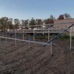 Metalowa konstrukcja wsporcza pod panele fotowoltaiczne, zamontowana na ziemi, w tle budynek gospodarczy i drzewa, widok o zmierzchu.