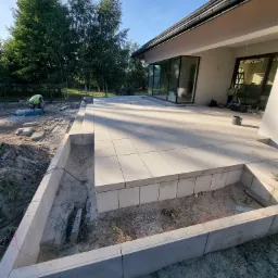 Jasny taras z płyt betonowych w trakcie budowy, widoczne betonowe obrzeża i przygotowane podłoże, pracownik w oddali obrabia kamień.
