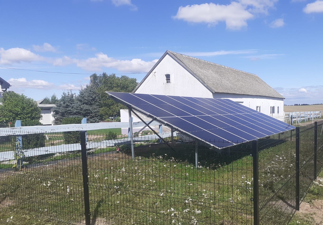 Instalacja fotowoltaiczna na metalowej konstrukcji, umieszczona na trawniku za ogrodzeniem z siatki, z białym budynkiem gospodarczym w tle.