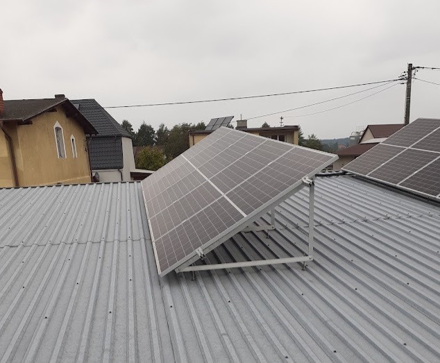 Panele fotowoltaiczne zamontowane na metalowym dachu budynku mieszkalnego, widoczne inne budynki w tle, pochmurne niebo.