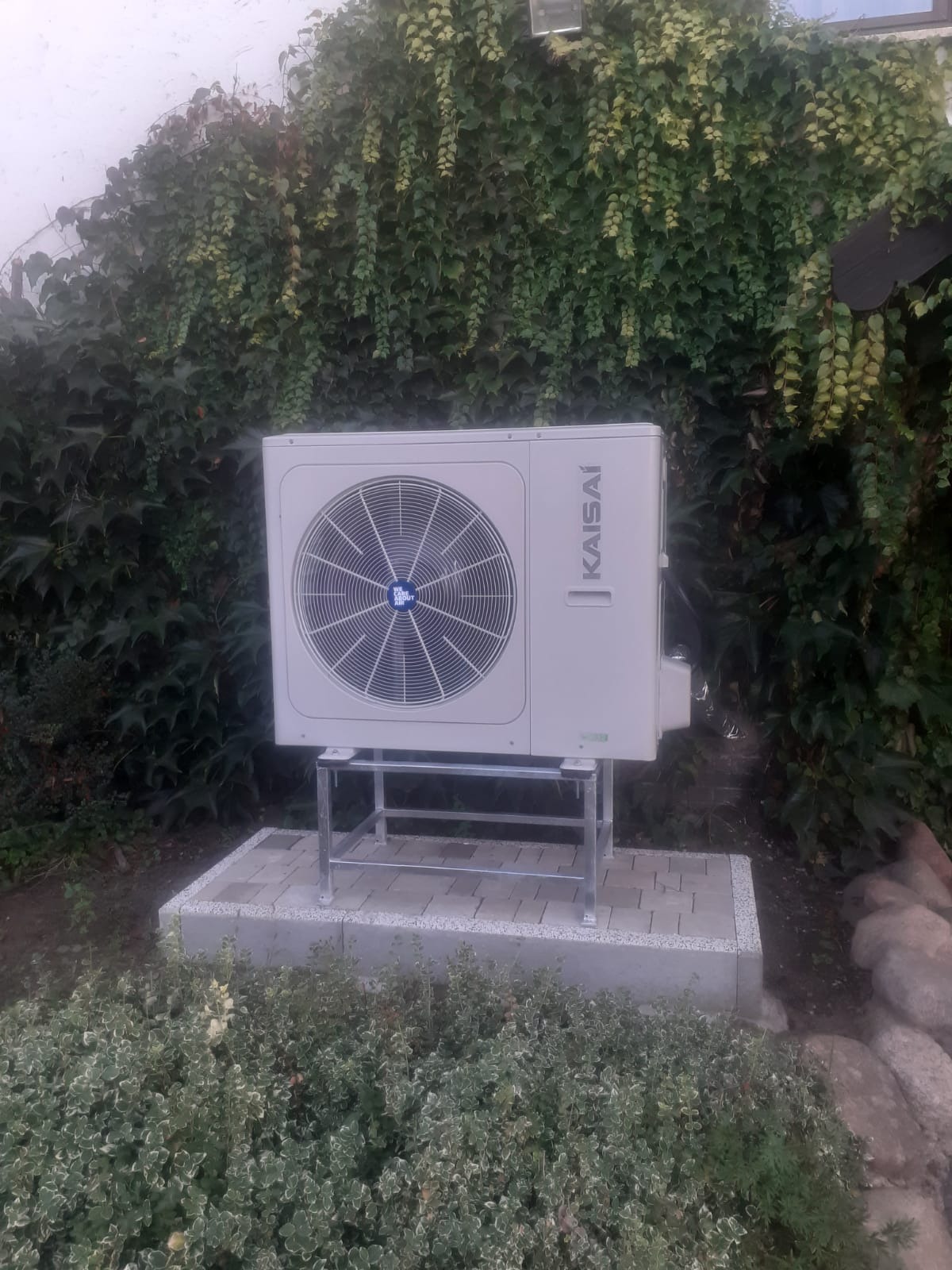 Zewnętrzna jednostka pompy ciepła marki Kaisai, zamontowana na metalowym stelażu na betonowym fundamencie, otoczona bujną zielenią bluszczu i krzewów.