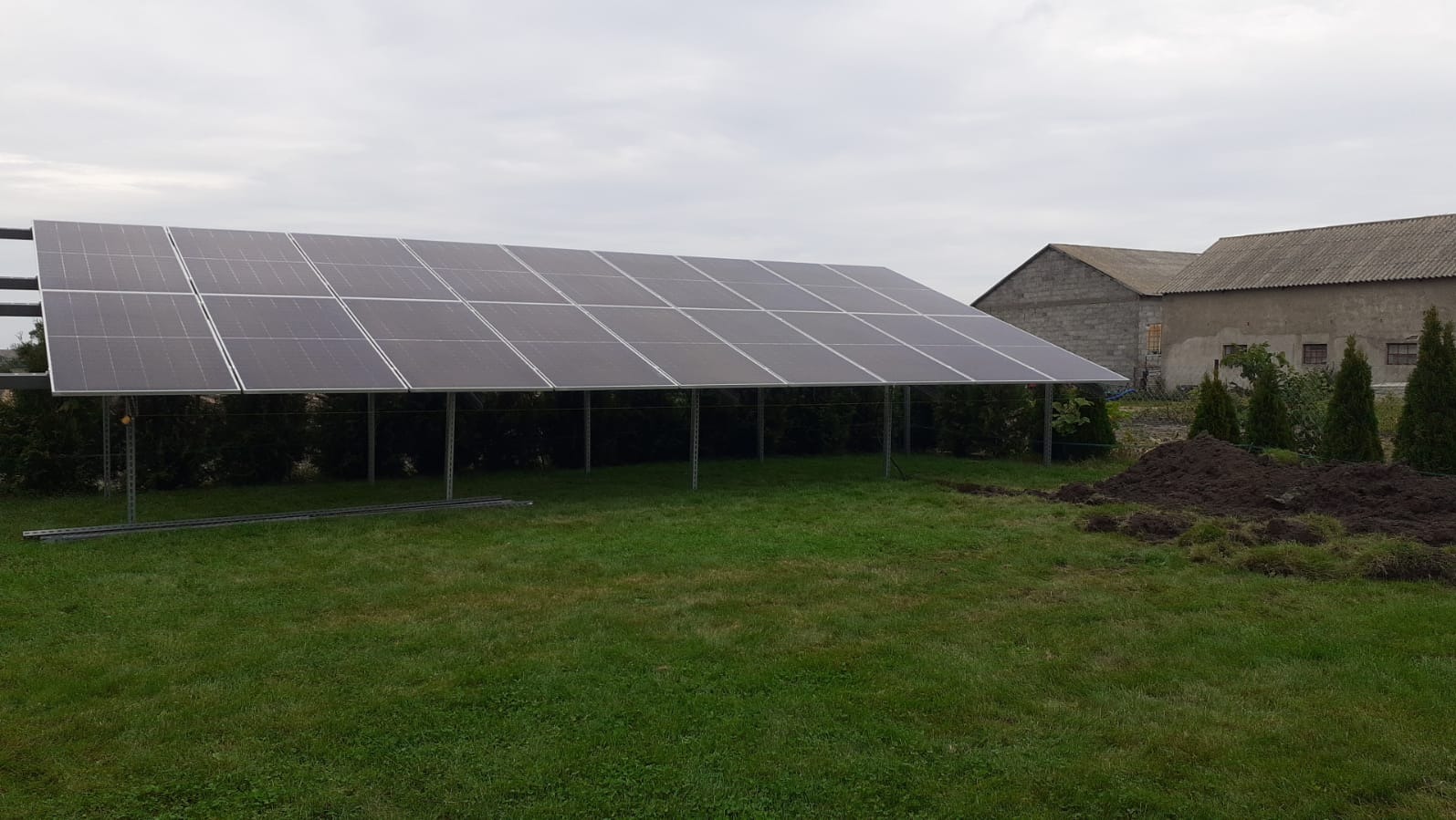 Montaż paneli słonecznych na metalowej konstrukcji w ogrodzie, widoczne przygotowane podłoże i budynki gospodarcze w tle.