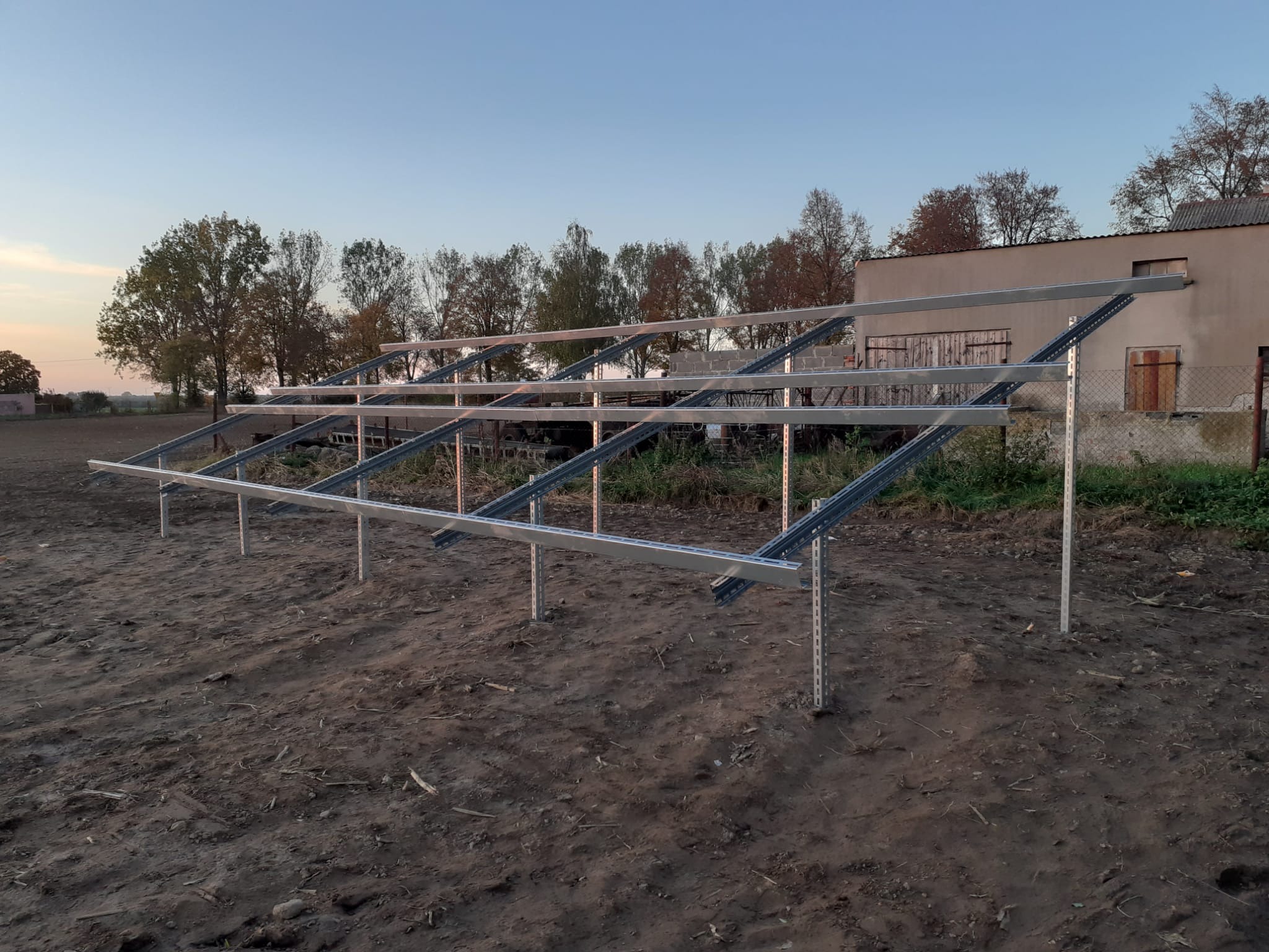 Metalowa konstrukcja wsporcza pod panele fotowoltaiczne, zamontowana na ziemi, w tle budynek gospodarczy i drzewa, widok o zmierzchu.