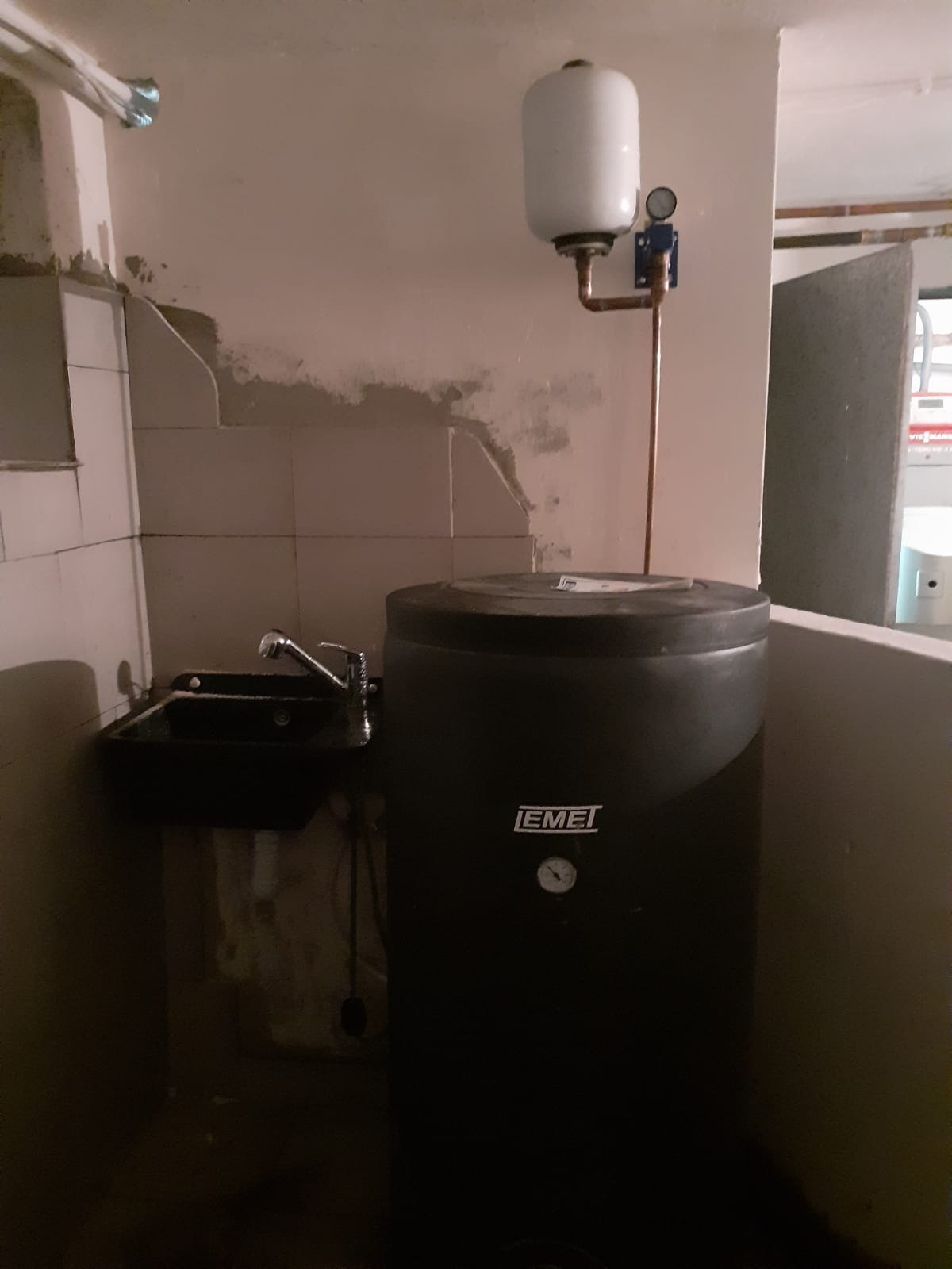 Czarny zasobnik ciepłej wody użytkowej marki EMEI z podłączonymi rurami miedzianymi i małym zbiornikiem wyrównawczym, umieszczony w pomieszczeniu z umywalką i częściowo wykończonymi ścianami.