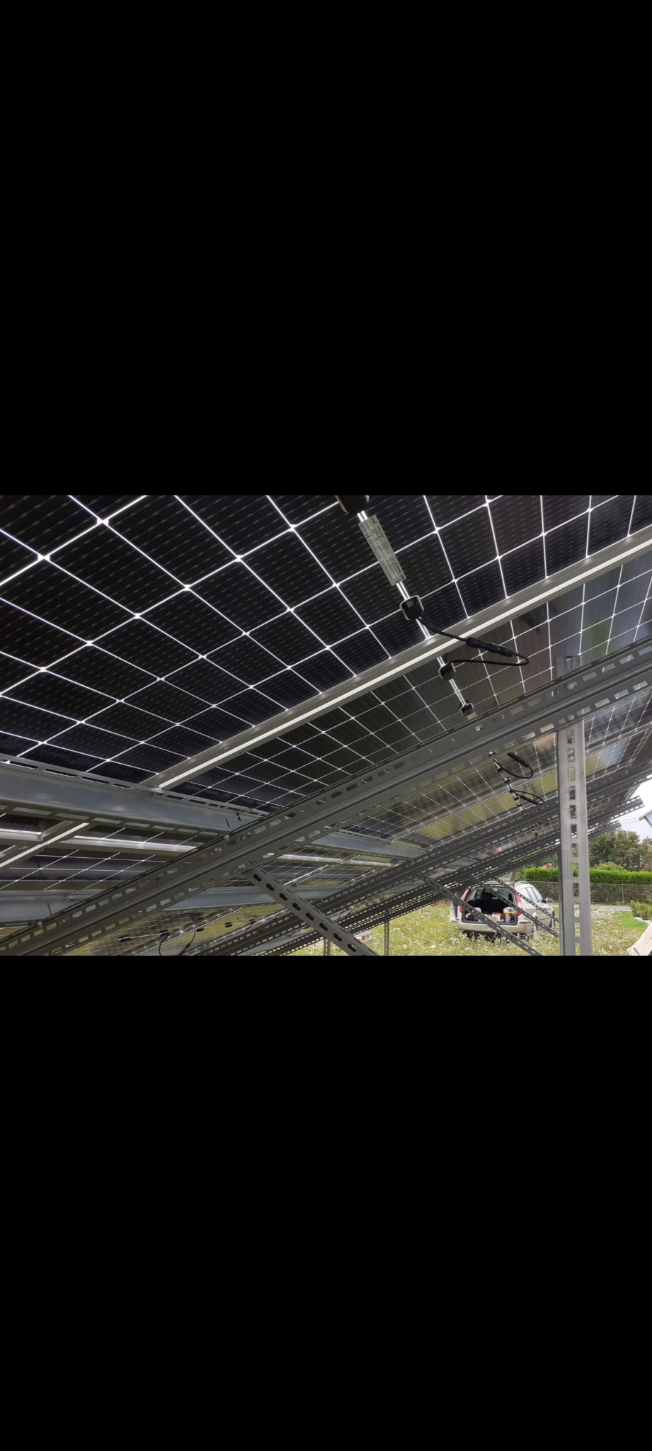 Montaż paneli fotowoltaicznych od spodu, widoczne metalowe konstrukcje wsporcze i częściowo samochód w tle. Instalacja w trakcie realizacji.