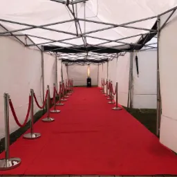 Czerwony dywan rozłożony wewnątrz białego namiotu z metalową konstrukcją w Wołominie, wyznaczony srebrnymi słupkami z czerwonym sznurem, na końcu widoczny grzejnik gazowy.