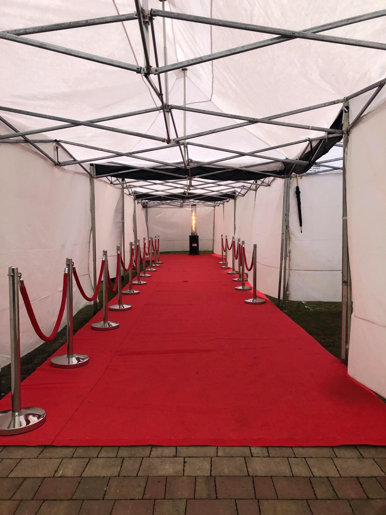 Czerwony dywan rozłożony wewnątrz białego namiotu z metalową konstrukcją w Wołominie, wyznaczony srebrnymi słupkami z czerwonym sznurem, na końcu widoczny grzejnik gazowy.