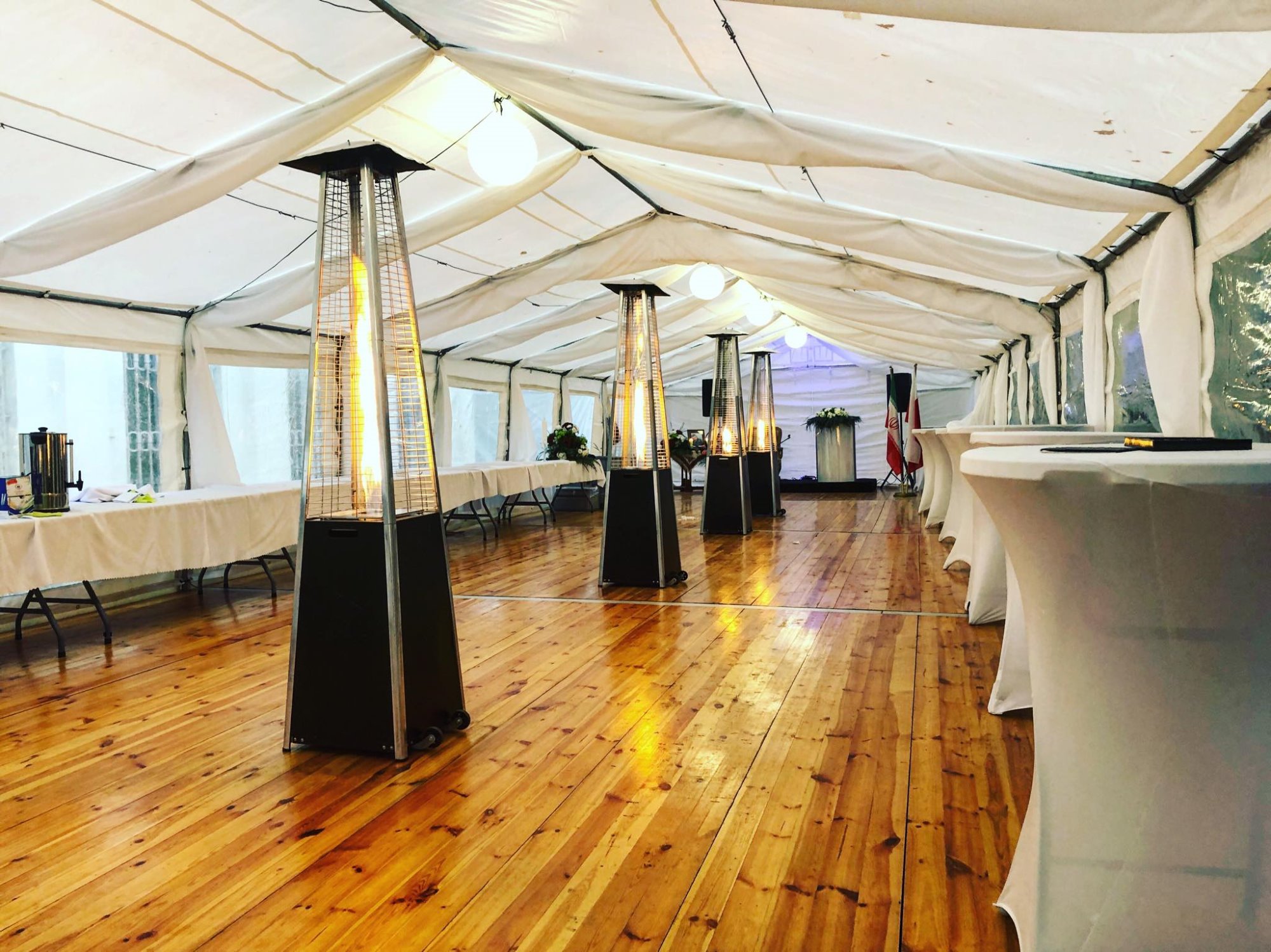 Wnętrze białego namiotu imprezowego z drewnianą podłogą, oświetlonego kulistymi lampami i ogrzewaczami gazowymi w kształcie kolumn. Białe stoły z obrusami ustawione wzdłuż ścian. W tle flaga Iranu.