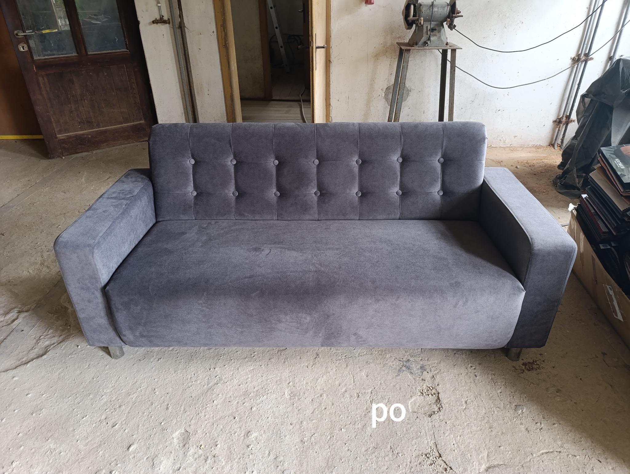 Szara sofa z guzikami na oparciu, sfotografowana w warsztacie z widocznymi narzędziami i materiałami.