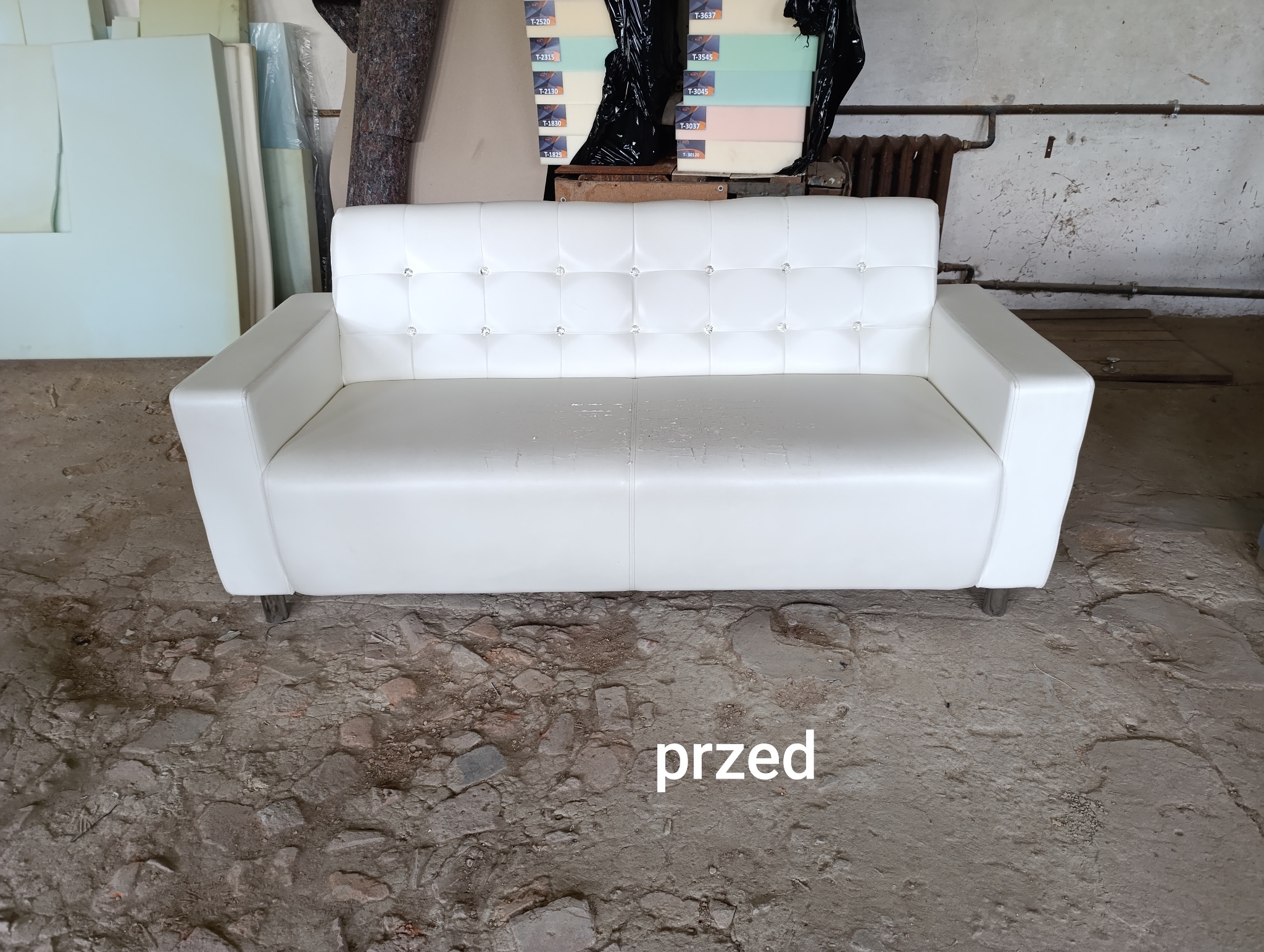 Biała, skórzana sofa z pikowanym oparciem i kryształkami, stojąca na betonowej podłodze w warsztacie tapicerskim, z widocznymi materiałami tapicerskimi w tle. Na podłodze widoczny napis 'przed'.