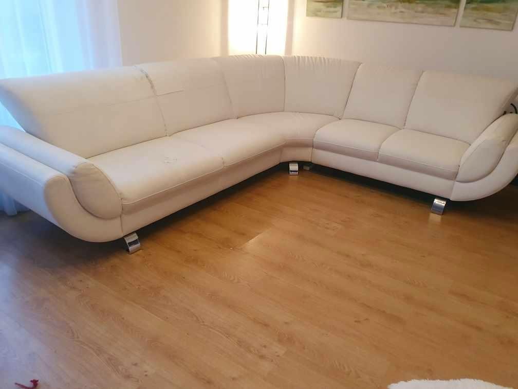 Jasny, skórzany narożnik z metalowymi nóżkami na drewnianej podłodze. Widoczne ślady użytkowania na siedzisku. W tle ściana z obrazami i lampą.