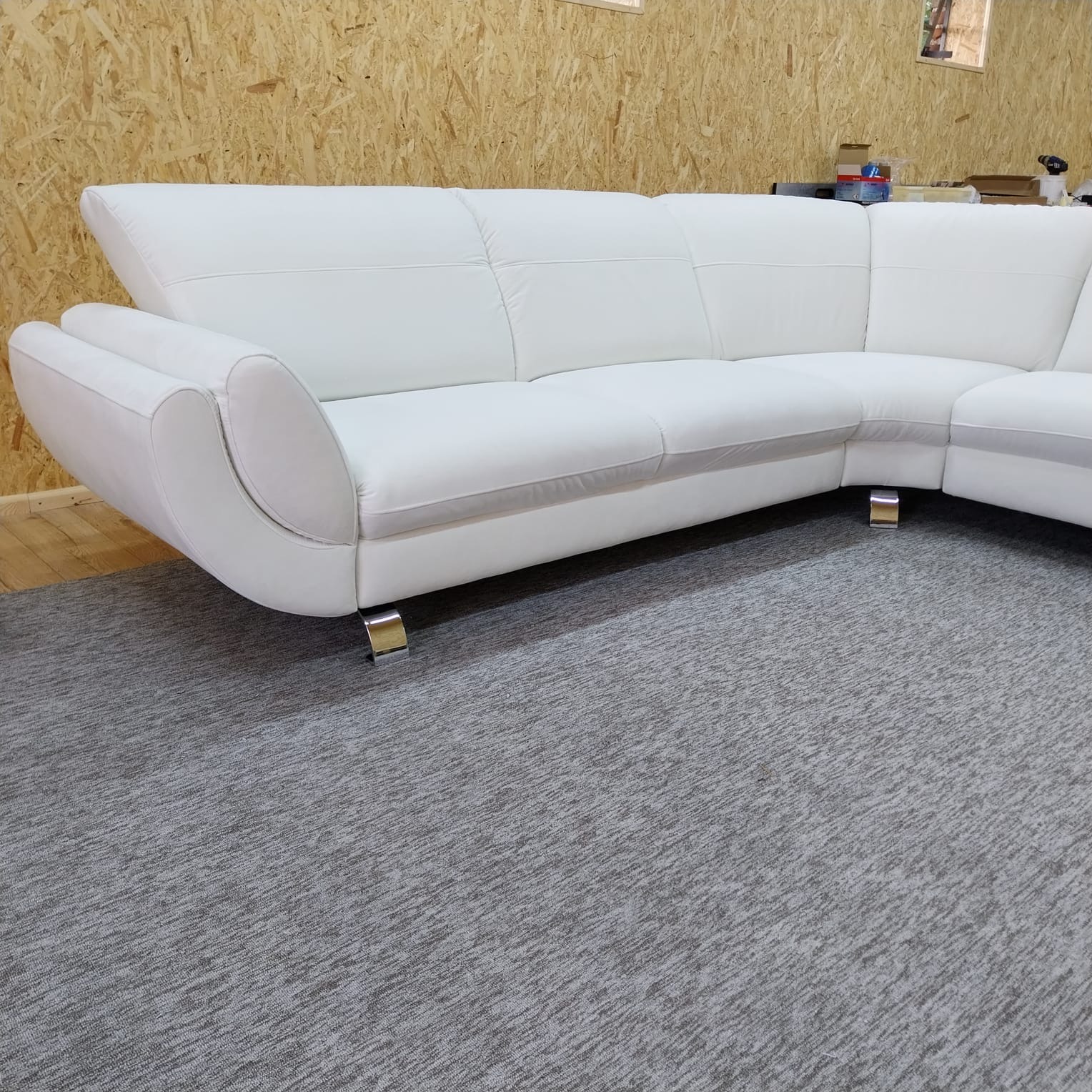 Biały, skórzany narożnik z chromowanymi nóżkami na szarym dywanie. Nowoczesny design, widoczne przeszycia i zaokrąglone podłokietniki. Tło stanowi ściana z płyt OSB.