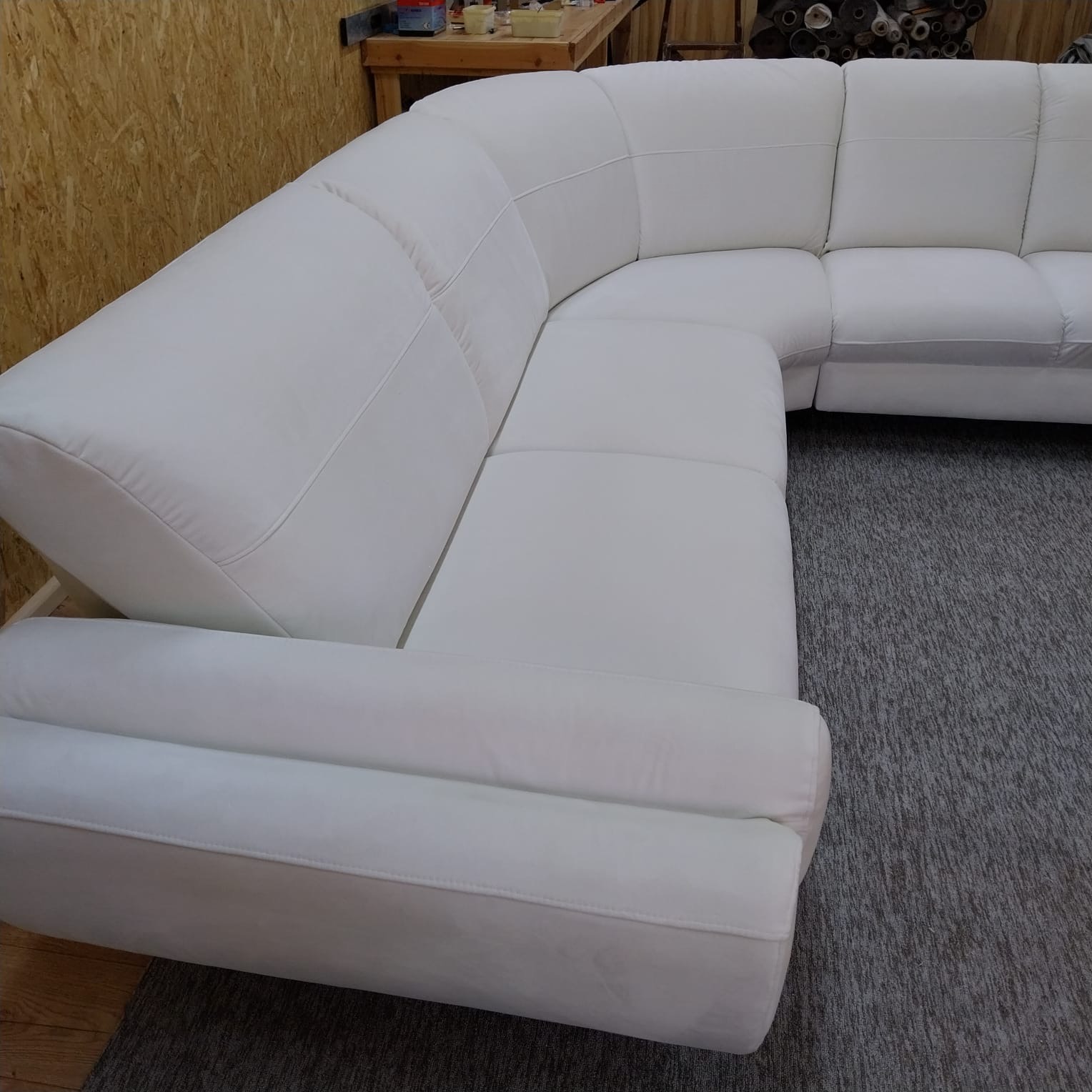 Biały, narożny zestaw wypoczynkowy z miękkim obiciem, widoczny z bliska na tle szarego dywanu i drewnianej ściany. Widoczne detale szwów i faktura materiału.