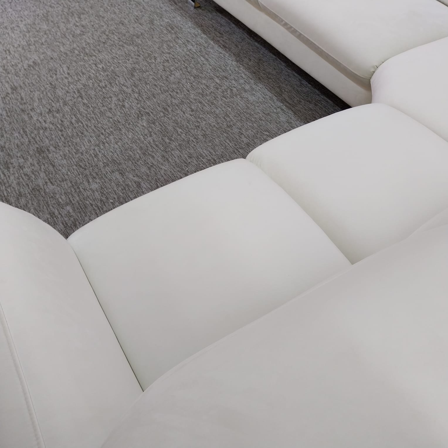 Fragment białej, modułowej sofy z miękkimi poduchami na tle szarego dywanu. Widok z góry, ukazujący detale tapicerki i fakturę materiałów.