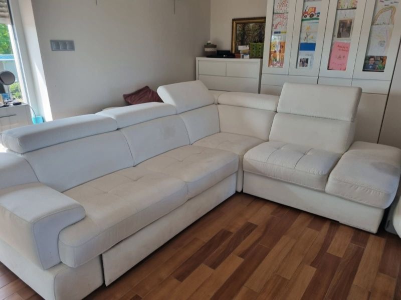 Biały, narożny zestaw wypoczynkowy z regulowanymi zagłówkami, widoczny na drewnianej podłodze w jasnym wnętrzu. W tle białe meble i dekoracje.