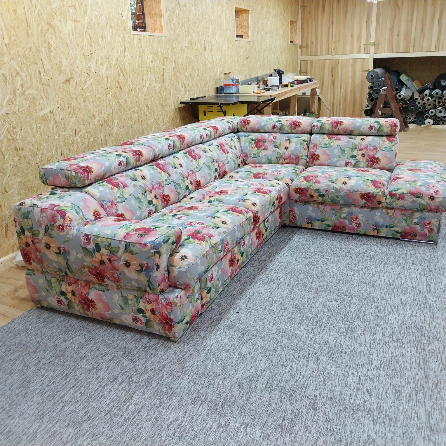 Narożnik po renowacji z kwiatowym wzorem, na tle warsztatu z narzędziami i rolkami materiałów. Jasne drewno na ścianach. Szary dywan. Ukończona usługa.