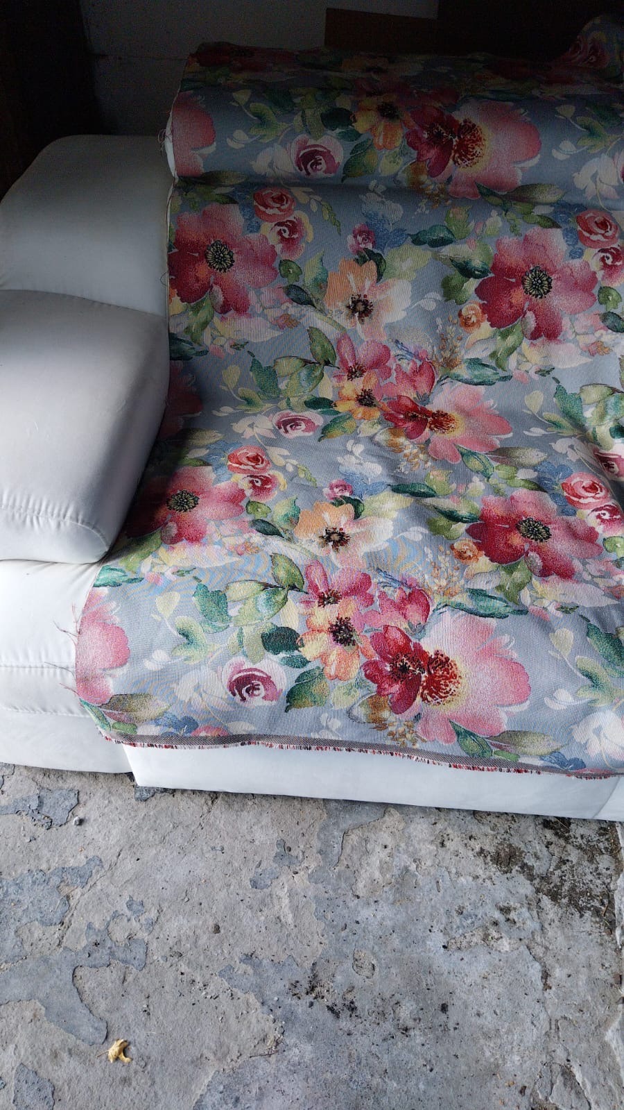 Biała sofa z nową, wzorzystą tkaniną w kwiaty, częściowo założoną. Widoczny fragment podłogi z szarą, popękaną powierzchnią. Ujęcie z góry, bliskie.