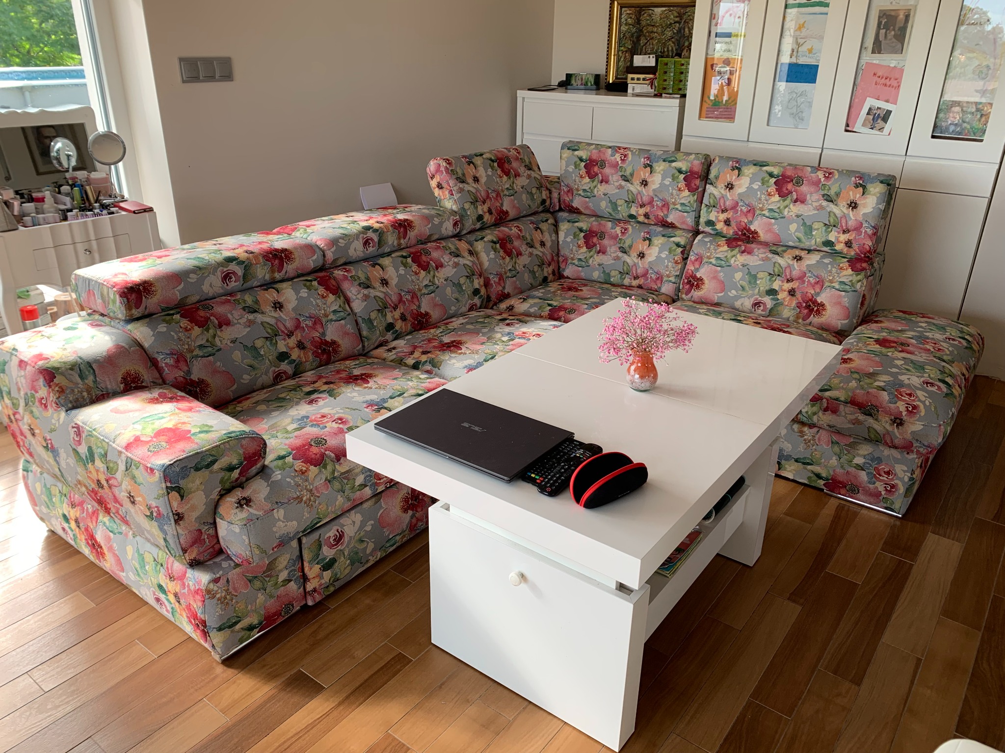 Narożnik po renowacji z kwiatowym wzorem, na białym stoliku laptop i pilot. Jasne wnętrze, drewniana podłoga. Widoczne detale tapicerki i nowoczesny design mebli.