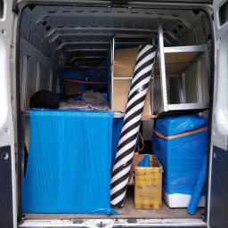 MoveHERO - Wnętrze furgonetki załadowane meblami i przedmiotami zabezpieczonymi folią, gotowymi do transportu. Widoczny zwinięty dywan w biało-czarne pasy oraz metalowy regał.