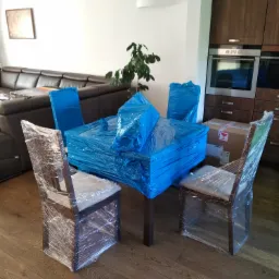 Stół i krzesła owinięte folią stretch oraz niebieską folią ochronną w trakcie przygotowań do transportu, widoczny narożnik skórzanej sofy w tle